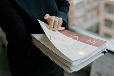 Closeup of someone’s hands holding a stack of U.S. patents. The top page reads “United States of America “ and “Patent” in gold lettering, among other smaller text. They are next to a window that looks down on a city street.