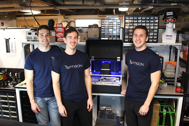 Brandon Motes, Dane deQuilettes, and Anthony Troupe in Optigon T-shirts stand in front of a bench bearing a glowing, suitcase-sized device